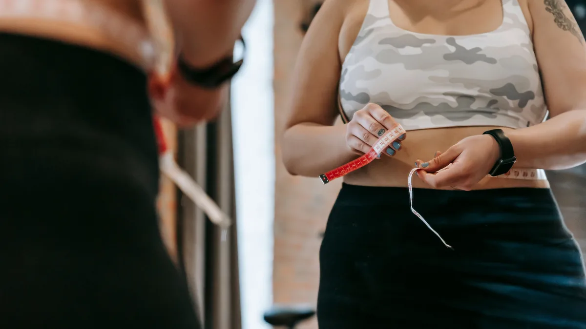 Woman in activewear measuring her waist with a tape measure in a gym.