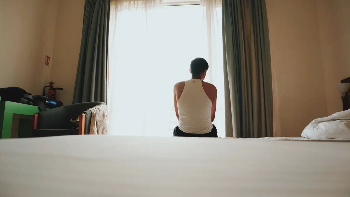 Man sitting on a bed facing a bright window.
