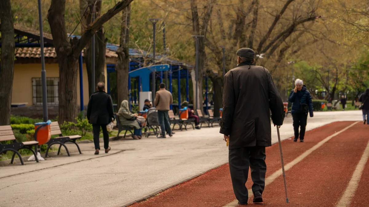 Older man walking with a cane along a tree-lined park path.