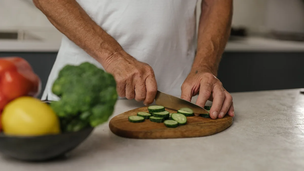 Adult slicing cucumber on a wooden board beside fresh vegetables.